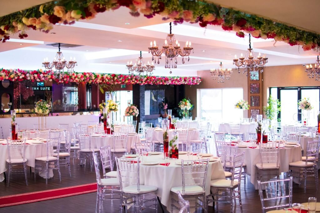 Decorated Asian wedding hall at The Maison Bradford with round tables, flower displays, chandeliers and a stage-ready layout for mehndi, nikah and walima events.