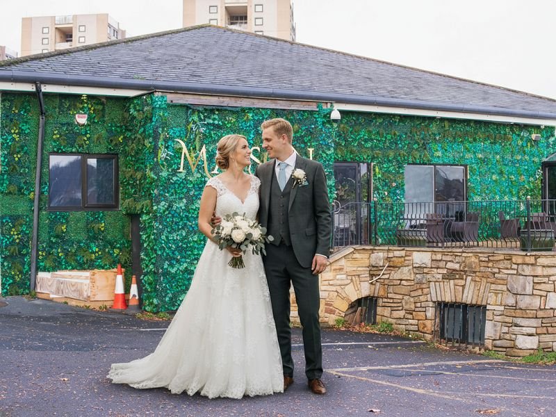 A happy bride and groom standing outside The Maison, a premier wedding reception venue in Bradford featuring a luxury green floral wall exterior.